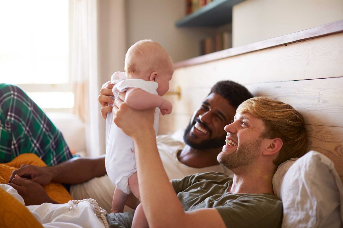 Two fathers sitting on their bed together admiring their newborn daughter.